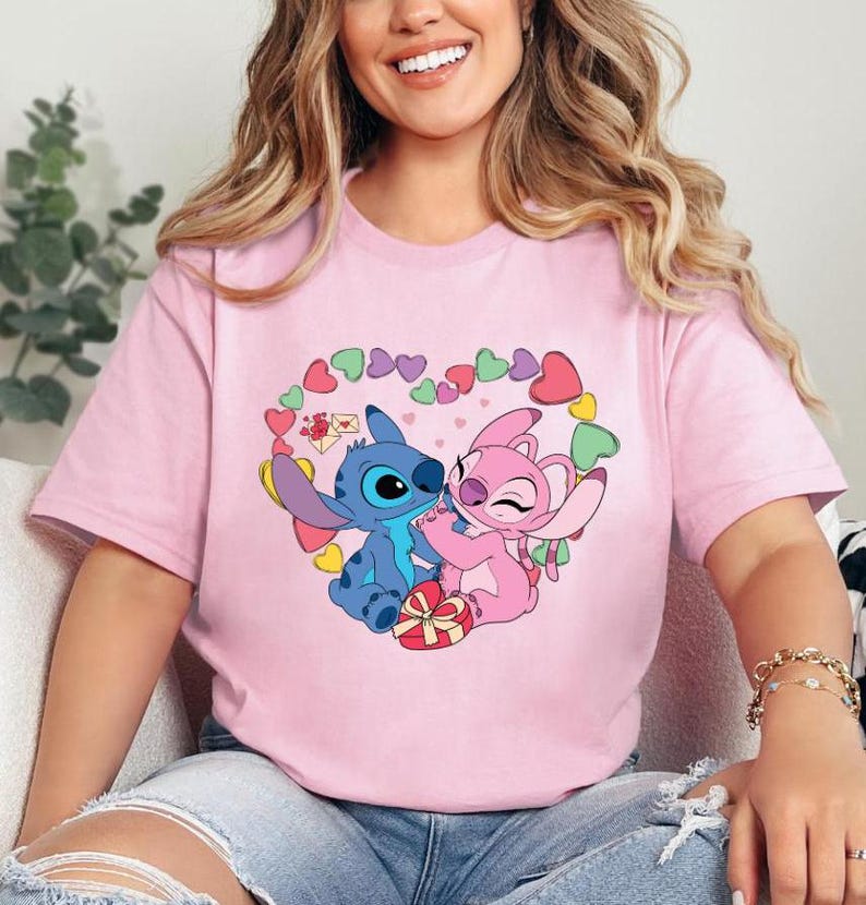 May include: Pink t-shirt featuring a graphic of Stitch and Angel from Lilo & Stitch. The graphic depicts Stitch and Angel embracing, surrounded by a heart shape made of pink and green hearts.