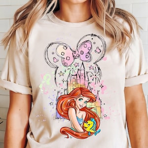 May include: A light beige t-shirt with a colorful graphic print of a cartoon mermaid holding a fish. The graphic also includes a castle, stars, and a pink bow.