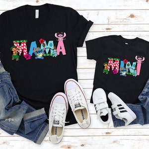 May include: Two black t-shirts with a graphic print of the words "MAMA" and "MINI" in colorful letters with Stitch and Lilo characters. The shirts are laid out on a white wooden surface with blue jeans and white sneakers.