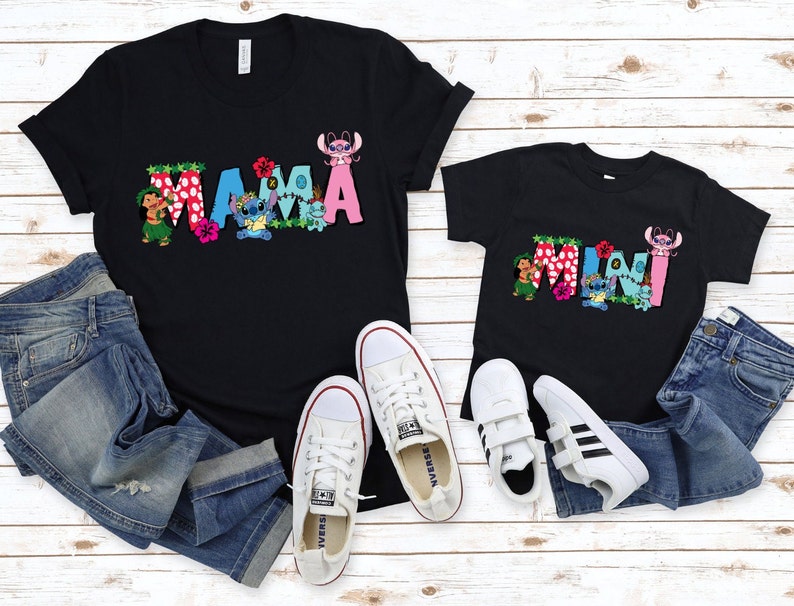 May include: Two black t-shirts with a graphic print of the words "MAMA" and "MINI" in colorful letters with Stitch and Lilo characters. The shirts are laid out on a white wooden surface with blue jeans and white sneakers.