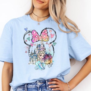 Puede incluir: Camiseta azul claro con un gr&aacute;fico de un lazo rosa y blanco, un castillo y una princesa con cabello rubio. El gr&aacute;fico est&aacute; rodeado de globos y fuegos artificiales coloridos. El texto "Elsa" est&aacute; escrito debajo del gr&aacute;fico.