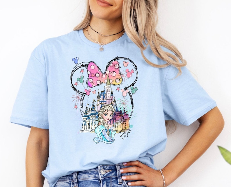 Puede incluir: Camiseta azul claro con un gr&aacute;fico de un lazo rosa y blanco, un castillo y una princesa con cabello rubio. El gr&aacute;fico est&aacute; rodeado de globos y fuegos artificiales coloridos. El texto "Elsa" est&aacute; escrito debajo del gr&aacute;fico.