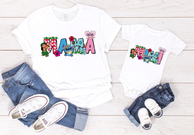 May include: White t-shirts with the words "MAMA" and "MINI" in colorful letters with cartoon characters. The shirts are on a wooden surface with blue jeans and white sneakers.
