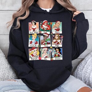 Puede incluir: Sudadera negra con un gr&aacute;fico de princesas Disney vestidas de Pap&aacute; Noel. Las princesas incluyen Blancanieves, Ariel, Aurora, Cenicienta, Tiana, Bella, Rapunzel, Elsa y Jasmine.