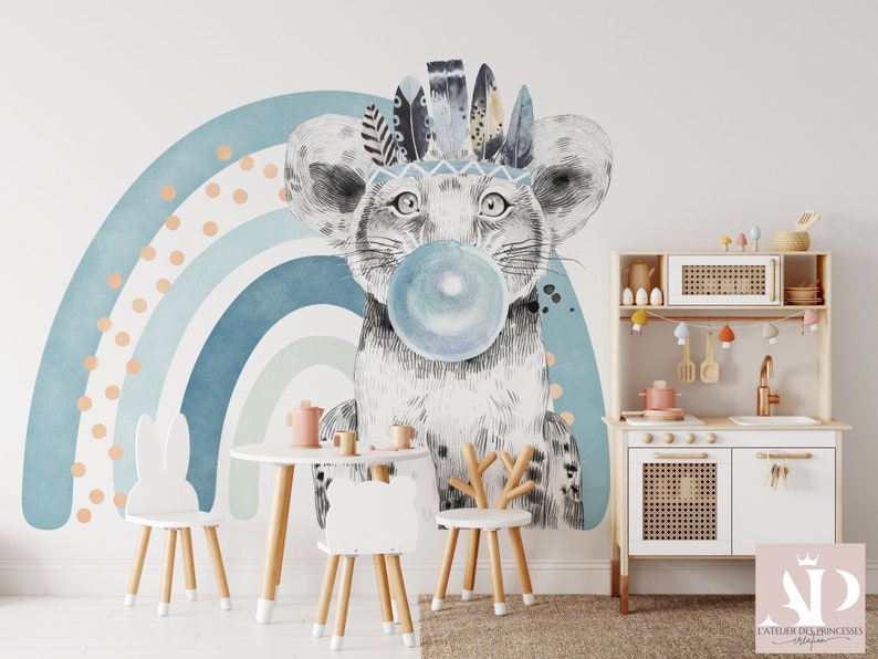 Puede incluir: Una cocina de juguete blanca con una encimera de madera y un armario blanco con una puerta de rat&aacute;n tejido. La cocina tiene un fregadero, una placa de cocci&oacute;n y un horno peque&ntilde;o. Hay decoraciones coloridas colgadas sobre la cocina. Una silla blanca de conejo y una silla blanca de ciervo est&aacute;n frente a la cocina. Un arco&iacute;ris azul y verde con lunares naranjas est&aacute; en la pared detr&aacute;s de la cocina.