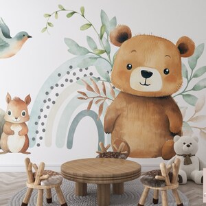 May include: A watercolor illustration of a brown bear, a squirrel, and a bluebird with a rainbow in the background. The illustration is on a white wall with two wooden chairs and a wooden table in front of it.