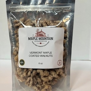 May include: A clear plastic bag filled with Vermont maple coated walnuts. The bag has a white label with the text "Maple Mountain Sugarhouse" and "Vermont Maple Coated Walnuts" with a maple leaf logo. The bag contains 8 ounces of walnuts.