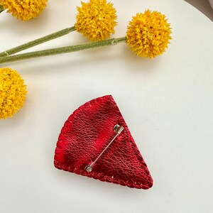 May include: A red leather brooch in the shape of a slice of pie with a silver pin on the back.
