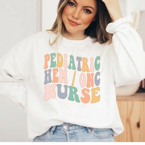 May include: White sweatshirt with the text "PEDIATRIC HEM/ONC NURSE" in colorful retro font.