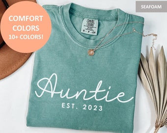 May include: A seafoam green t-shirt with the text "Auntie Est. 2023" in white. The shirt is folded and has a tag that says "Comfort Colors".