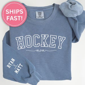 K&ouml;nnte beinhalten: Blauer Jeans-Sweatshirt mit dem Text "HOCKEY MOM" in Wei&szlig; auf der Vorderseite und dem Text "RYAN & MATT" mit Hockeyschl&auml;gern auf dem linken &Auml;rmel.