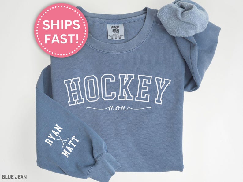 K&ouml;nnte beinhalten: Blauer Jeans-Sweatshirt mit dem Text "HOCKEY MOM" in Wei&szlig; auf der Vorderseite und dem Text "RYAN & MATT" mit Hockeyschl&auml;gern auf dem linken &Auml;rmel.