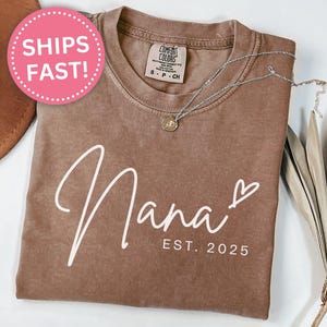 May include: A brown t-shirt with the text "Nana Est. 2025" in white lettering. The shirt has a distressed look and is made of a soft, comfortable fabric.
