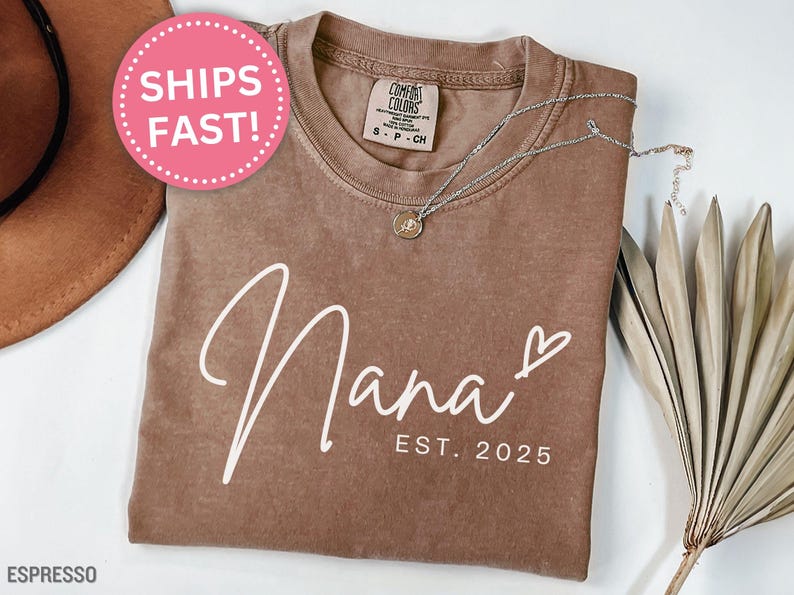 May include: A brown t-shirt with the text "Nana Est. 2025" in white lettering. The shirt has a distressed look and is made of a soft, comfortable fabric.