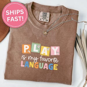 May include: An espresso-colored short-sleeve t-shirt featuring a graphic with the text PLAY IS MY FAVORITE LANGUAGE. The word PLAY is written in colorful blocks with American Sign Language hand signs, and the remaining text is in white and colorful letters.