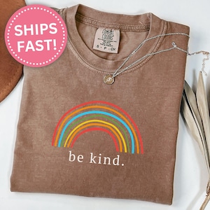 Può includere: T-shirt color espresso con grafica arcobaleno e la scritta "be kind". L'arcobaleno presenta strisce rosse, arancioni, gialle, verdi e blu. Una collana d'argento con un piccolo ciondolo è sulla maglietta.