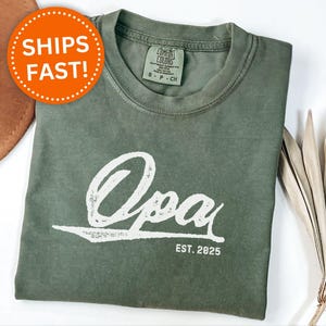 May include: A moss green t-shirt with the word "Opa" in a white, stylized font. The text "EST. 2025" is below the word "Opa". The shirt is folded and has a tag on the collar. A brown hat is partially visible in the background.