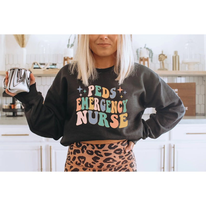 May include: Black sweatshirt with the text "PEDS EMERGENCY NURSE" in colorful retro font. The text is surrounded by small stars.