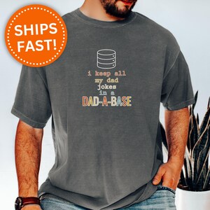 May include: Dark heather gray t-shirt with the text "i keep all my dad jokes in a DAD-A-BASE" in a colorful font. The shirt also has a graphic of a database server. An orange circle with the words "SHIPS FAST!" is in the upper left.