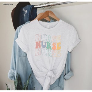 Pu&ograve; includere: Una maglietta bianca con la parola "NURSE" ripetuta tre volte in un font retr&ograve;. Ogni parola &egrave; di un colore pastello diverso: rosa, blu, verde e giallo.