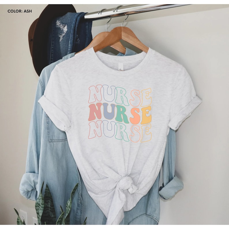 Pu&ograve; includere: Una maglietta bianca con la parola "NURSE" ripetuta tre volte in un font retr&ograve;. Ogni parola &egrave; di un colore pastello diverso: rosa, blu, verde e giallo.