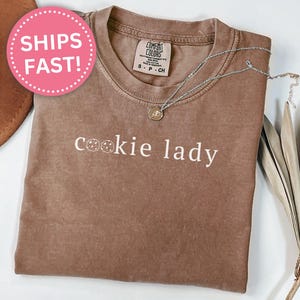 May include: A brown t-shirt with the words "cookie lady" in white script. The shirt has a crew neck and short sleeves. A silver necklace with a pendant is draped on the shirt. A pink sticker with the words "SHIPS FAST!" is in the upper left corner.