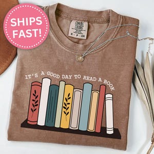 Può includere: T-shirt color espresso con la frase "It's a good day to read a book" e una grafica di libri colorati su uno scaffale. La maglietta ha un girocollo e maniche corte. Una collana d'argento con un ciondolo è sulla maglietta.