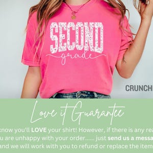 May include: A bright pink t-shirt with the words "SECOND grade" in a white floral design. The shirt is a crew neck style and is made of a soft material. The brand name "CRUNCHBERRY" is printed on the bottom right.