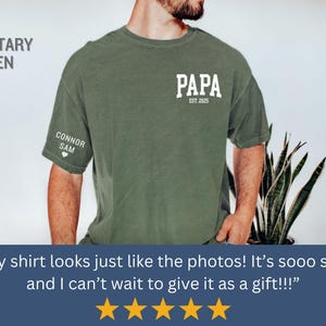 Op de afbeelding: Militairgroen t-shirt met het woord "PAPA" en "EST. 2025" in het wit gedrukt. De linkermouw heeft "CONNOR SAM" en een hart. Het shirt is een comfortabele, casual stijl.