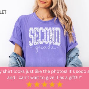 May include: A violet-colored t-shirt with the words "SECOND grade" in white, floral-filled letters. The shirt has rolled-up sleeves and is being worn. The word "VIOLET" is in the upper left corner.