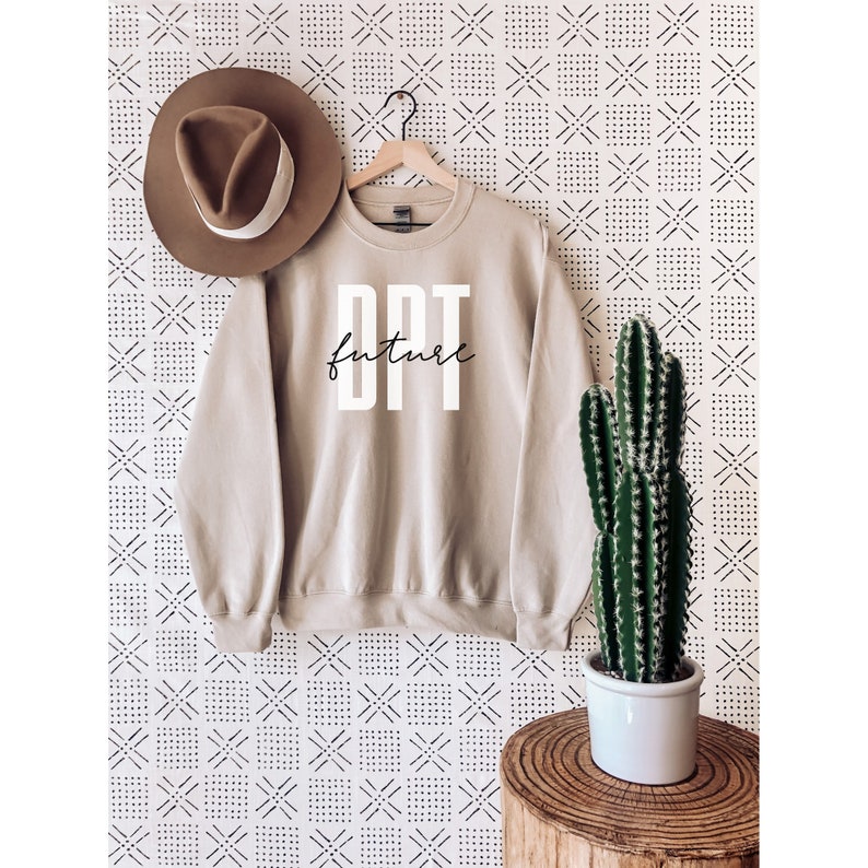 Pu&ograve; includere: Una felpa beige con la scritta "DPT future" stampata in bianco sul davanti. La felpa &egrave; appesa a un appendiabiti su uno sfondo bianco con un motivo geometrico bianco e nero. Nell'immagine sono visibili anche un cappello marrone e una pianta di cactus.