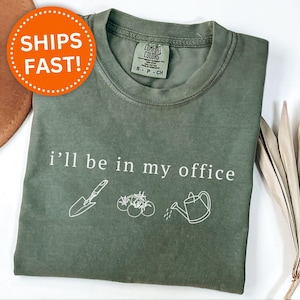 May include: A moss green t-shirt featuring the phrase "i'll be in my office" and gardening-related illustrations. The design includes a trowel, tomatoes, and a watering can. The shirt has a crew neck and short sleeves.