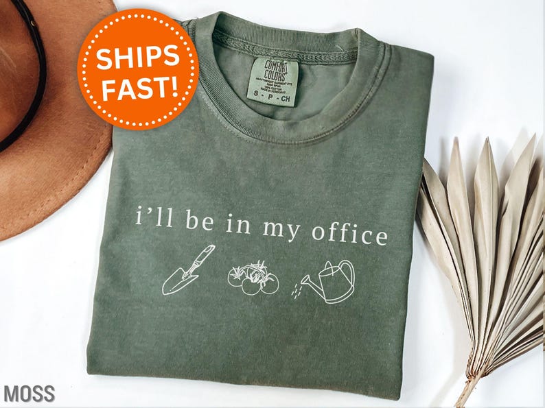 May include: A moss green t-shirt featuring the phrase "i'll be in my office" and gardening-related illustrations. The design includes a trowel, tomatoes, and a watering can. The shirt has a crew neck and short sleeves.