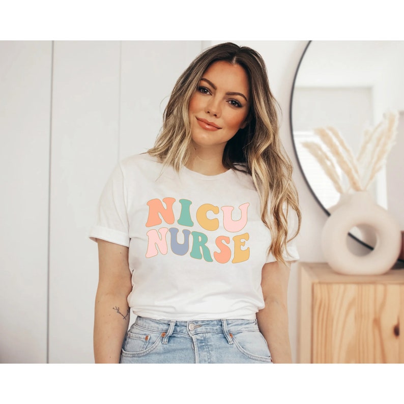 Pode incluir: Uma camiseta branca com as palavras "NICU NURSE" em letras retr&ocirc; coloridas.