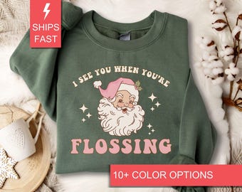 Op de afbeelding: Groen sweatshirt met een cartoon afbeelding van de kerstman en de tekst "I see you when you're flossing".