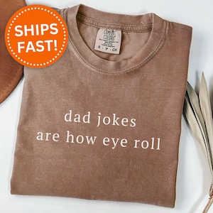 May include: Espresso-colored t-shirt with the text "dad jokes are how eye roll" in white. The shirt has a crew neck and a Comfort Colors tag. An orange circle with the words "SHIPS FAST!" is in the upper left corner.