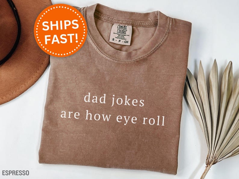 May include: Espresso-colored t-shirt with the text "dad jokes are how eye roll" in white. The shirt has a crew neck and a Comfort Colors tag. An orange circle with the words "SHIPS FAST!" is in the upper left corner.