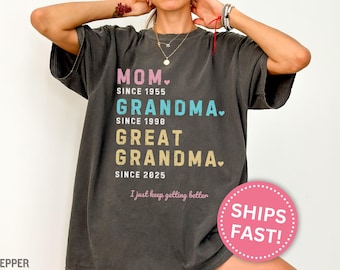 May include: A dark gray t-shirt with the text "MOM. SINCE 1955 GRANDMA. SINCE 1998 GREAT GRANDMA. SINCE 2025 I just keep getting better"