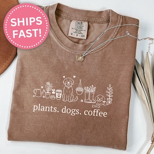 May include: Espresso-colored t-shirt with a white graphic of plants, dogs, and coffee cups. The text "plants. dogs. coffee" is printed below the graphic. A silver necklace is draped on the shirt.