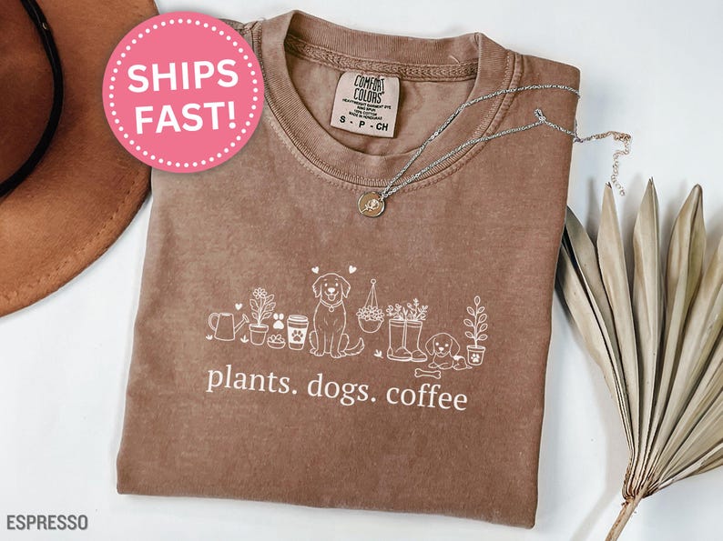 May include: Espresso-colored t-shirt with a white graphic of plants, dogs, and coffee cups. The text "plants. dogs. coffee" is printed below the graphic. A silver necklace is draped on the shirt.