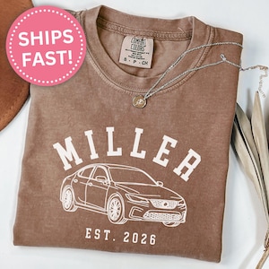 Può includere: T-shirt color espresso con il nome "MILLER" sopra una grafica di auto e la scritta "EST. 2026". Una collana d'argento con un ciondolo è sulla maglietta. La maglietta è piegata su una superficie bianca.