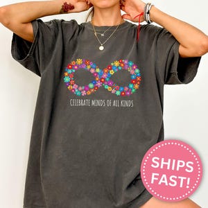 Può includere: T-shirt grigio scuro con un simbolo infinito floreale multicolore e la scritta "Celebrate Minds of All Kinds". La maglietta ha una vestibilità ampia.