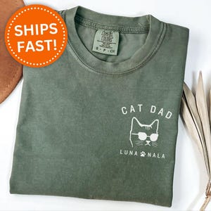 May include: Moss green t-shirt with a white graphic of a cat wearing sunglasses and the words "CAT DAD" and "LUNA NALA". The shirt is made of a soft material and has a crew neck. The shirt is a size small.