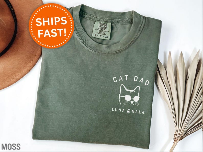 May include: Moss green t-shirt with a white graphic of a cat wearing sunglasses and the words "CAT DAD" and "LUNA NALA". The shirt is made of a soft material and has a crew neck. The shirt is a size small.