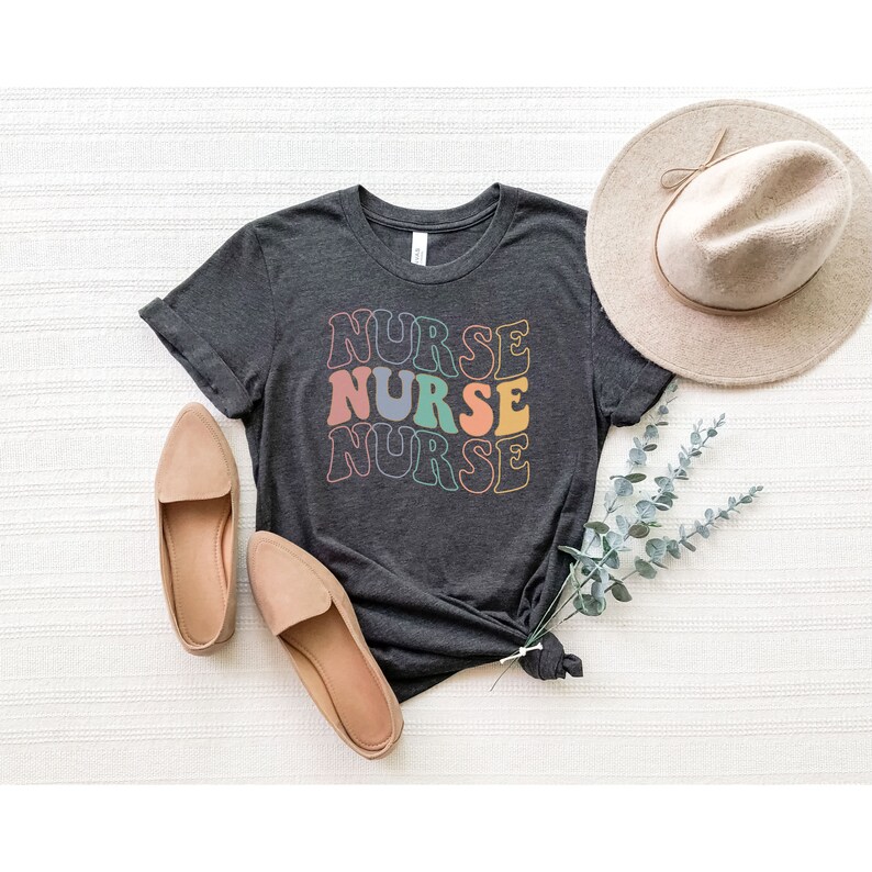 Pu&ograve; includere: Una maglietta grigio scuro con la parola "NURSE" ripetuta tre volte in un font retr&ograve;. Ogni parola &egrave; di un colore diverso, tra cui rosa, arancione, verde acqua e grigio.