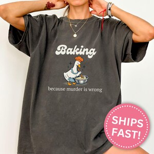Può includere: T-shirt grigio scuro con la scritta "Baking" in un font retrò sopra un'oca dei cartoni animati con grembiule e cappello da chef, che mescola gli ingredienti in una ciotola. Il testo "because murder is wrong" è sotto.