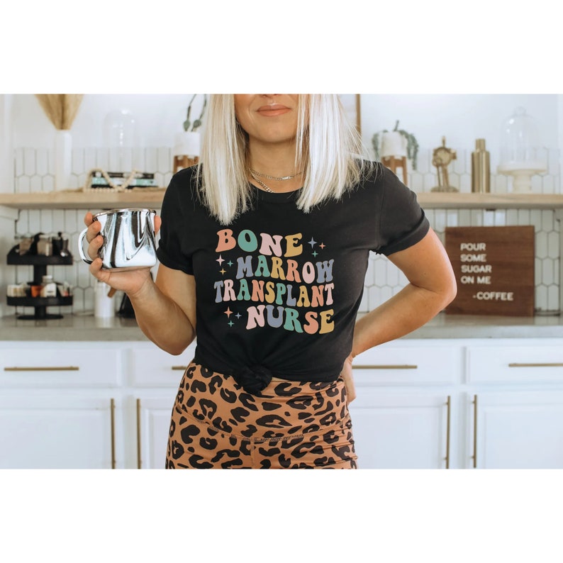 Może przedstawiać: Czarny T-shirt z napisem "BONE MARROW TRANSPLANT NURSE" w kolorowej czcionce retro. Tekst jest otoczony małymi gwiazdkami.