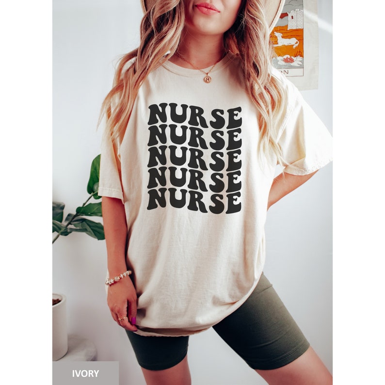 May include: A person wearing a light beige t-shirt with the word "NURSE" repeated five times in a retro font. The shirt is oversized and has short sleeves. The person is wearing dark green cycling shorts.