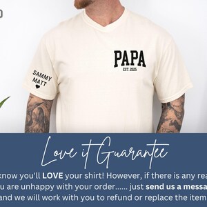 Op de afbeelding: Zandkleurig T-shirt met het woord "PAPA" in vette zwarte letters en "EST. 2025" eronder. Op de linkermouw staan de namen "SAMMY" en "MATT" met een hartje. Het shirt maakt deel uit van een kledingcollectie.