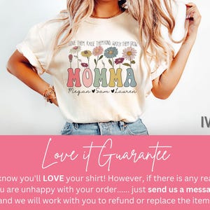 May include: A white t-shirt with a floral design and the text "LOVE THEM RAISE THEM KIND, WATCH THEM GROW MOMMA Megan Sam Lauren" in colorful letters. The shirt is shown on a model wearing blue jeans. The background is a pink and white color.